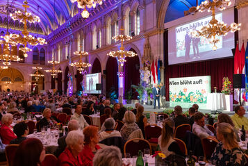 Blick in den vollbesetzten großen Festsaal des Wiener Rathauses. An den Tischen sitzen Menschen, im Vordergrund eine Bühne auf der gerade jemand von Karl Ploberger interviewt wird. Alles ist prächtig mit Blumen in den unterschildichsten Farben geschmückt.