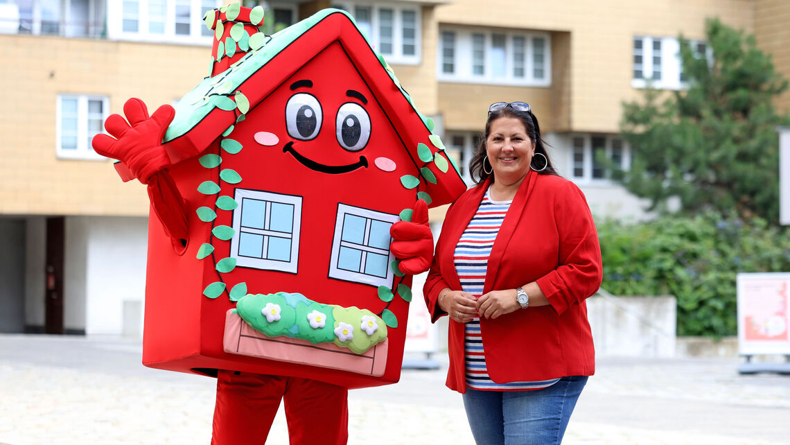 Vizebürgermeisterin und Wohnbaustadträtin Kathrin Gaál posiert neben dem Maskottchen für den Wiener Wohnbau, der Wohna Lisa. Das Kostüm der Wohna Lisa besteht aus einem roten Haus mit viel Blumenschmuck an den Fenstern, Augen, Mund, Armen und Beinen.