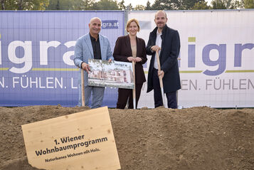 Drei Personen stehen auf einer Baustelle hinter einem Schild mit der Aufschrift '1. Wiener Wohnbaumprogramm Naturnahes Wohnen mit Holz' und halten ein Plakat vor einem Bauzaun mit dem Schriftzug 'migra.at'