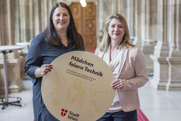 Vizebürgermeisterin und Frauenstadträtin Kathrin Gaál und Vizebürgermeisterin und Bildungsstadträtin Bettina Emmerling präsentieren die „Mädchen feiern Technik“-Workshops mit einem runden Schild, das von beiden gemeinsam gehaltne wird. Das Bild ist in den Arkadengängen des Rathauses aufgenommen worden. Kathrin Gaál trägt ein dunkelblaues Kleid, Bettina Emmerling eine dunkelblaue Hoseein beiges Oberteil mit Schalkragen und einen blassrosa Blazer darüber. Beide tragen ihre Haare offen.