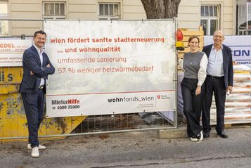Drei Personen stehen vor einem Bauzaun mit einem Banner, das Informationen zur Stadtentwicklung und Wohnqualität in Wien zeigt