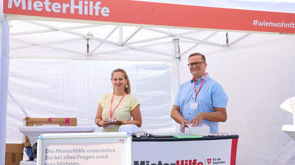 Zwei Personen stehen lächelnd an einem Informationsstand mit dem Schriftzug 'MieterHilfe' unter einem weißen Zelt.
