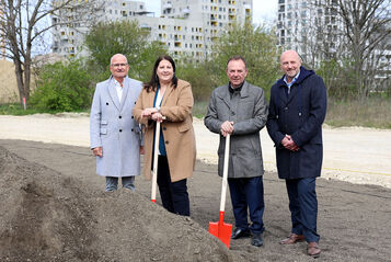 Dieter Groschopf, stellvertretender Geschäftsführer des wohnfonds_wien, Vizebürgermeisterin und Wohnbaustadträtin Kathrin Gaál, Bezirksvorsteher Gerald Bischof und WIGEBA-Vorstand Paul Steurer stehen an Spaten lehnend bei einem Sandhaufen. Die Aufnahme wurden offenbar zu einer kälteren Jahreszeit gemacht, da alle Protagonisten mit warmer Kleidung zu sehen sind.