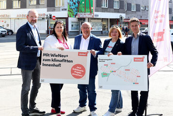 Drei Männer und zwei Frauen halten zwei Schilder mit den Inhalten "Mit Wien Neu+ zum klimafitten Innenhof" und einem Plan des Grätzls. Die fünf Personen sehen freundlich in die Kamera. Im Hintergrund ist eine Straßenkreuzung mit Schienen und vorbeifahrenden Autos zu erkennen.