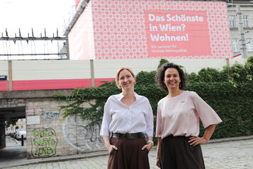 Zwei Frauen stehen vor einer mit Graffiti besprühten Mauer und einem Plakat mit der Aufschrift 'Das Schönste in Wien? Wohnen!'
