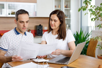 2 Personen sitzen an einem Holztisch und haben ein Notebook vor sich stehen. Zusätzlich hantieren sie mit einigen Zetteln und schauen diese konzentriert an. Im Hintergrund ist eine Küche erkennbar.