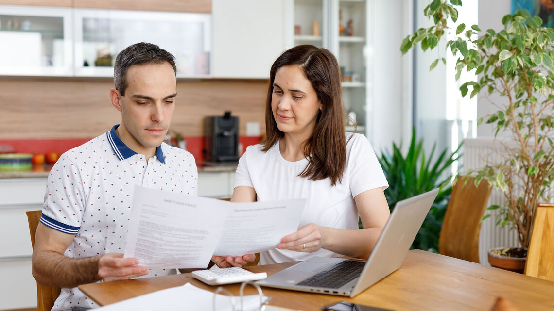 2 Personen sitzen an einem Holztisch und haben ein Notebook vor sich stehen. Zusätzlich hantieren sie mit einigen Zetteln und schauen diese konzentriert an. Im Hintergrund ist eine Küche erkennbar.