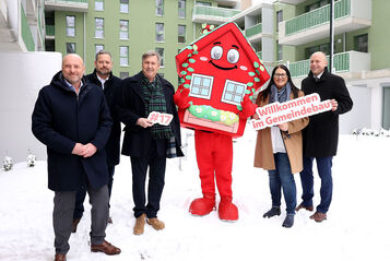 5 Personen stehen in winterlicher Business-Bekleidung im verschneiten Innenhof einer Neubauanlage. Ihnen zur Seite wurde wurde Wohna Lisa, das Maskottchen des geförderten Wohnbaus in Wien. Es ist eine als Haus verleidete rote Figur in Form eines Hauses mit Satteldach, großen Augen und einem freundlichen Lächeln.