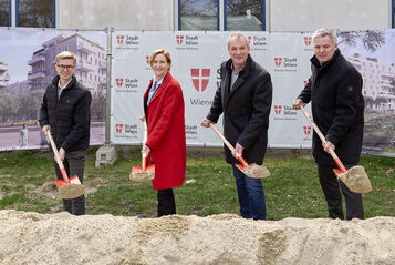 4 Personen stehen mit Spaten mit Sand darauf hinter einem Sandhaufen und lächeln in die Kamera. I Hintergrund ist ein Bauzaun mit Transparenten auf denen Visualisierungen des Gemeindebau NEU und das Logo von Stadt Wien/Wiener Wohnen zu erkennen ist.