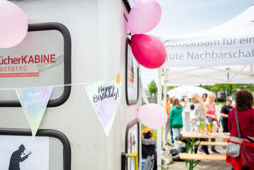 Links im Vordergrund ist eine Telefonzelle zu erkennen, die zu einer Bücherkabine umgebaut wurde. Sie ist mit Luftballons und mit einer Wimpel-Girlande geschmückt, auf der Happy Birthday steht. Im Hintergrund sind Menschen bei und unter einem wohnpartner-Pavillionzelt zu erkennen. auf einem Heurigentisch stehen Saftboxen. 