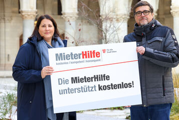 Kathrin Gaál und Christian Bartok stehen im Innenhof des Rathauses und halten gemeinsam eine Tafel auf der das Logo der MieterHilfe abgebildet ist und folgender Spruch zu lesen ist: "Die MieterHilfe unterstützt kostenlos!". Die beiden sind winterlich gekleidet.