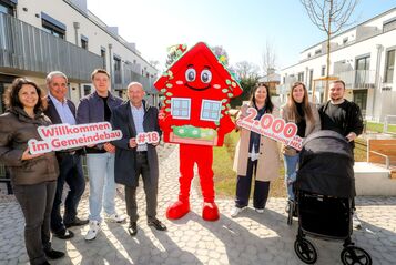 Sieben Personen stehen im Innenhof einer Wohnhausanlage. Die Anlage ist neu und hat drei Geschoße. In der Mitte der Gruppe steht das Maskottchen des Wiener Wohnbaus, Wohna Lisa. Sie ist ein rotes freundlich dreinblickendes Haus mit Satteldach auf zwei Beinen. Die Personen halten die Schriftzüge "Willkommen im Gemeindebau", "#18" und "2.000ste Gemeindewohnung NEU" in der Hand. Vor zwei Personen steht ein Kinderwagen. 