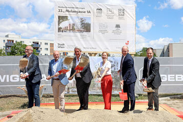 Sechs Personen mit Spaten stehen auf einer Baustelle vor einem großen Schild und schaufeln Erde in eine Sandkiste