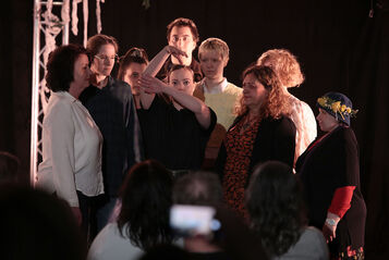 Mehrere Personen stehen knapp beisammen und blicken auf eine in der Mitte platzierte Person die mit ihren Armen die Äste eines Baumes darstellt. Der Hintergrund ist schwarz, im Vordergrund erkennt man Personen sitzend von hinten, die auf die Personengruppe schauen. 