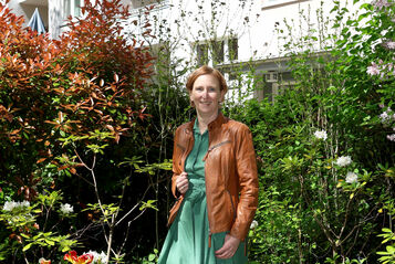 Frau in grünem Kleid und brauner Lederjacke steht in einem üppigen Garten vor einem mehrstöckigen Wohnhaus