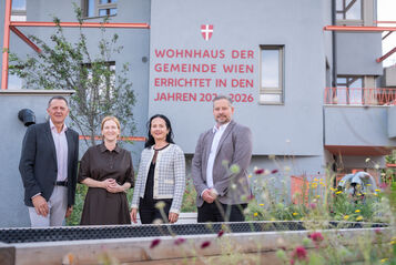 Vier Personen stehen vor einem modernen Wohnhaus mit der Aufschrift 'Wohnhaus der Gemeinde Wien errichtet in den Jahren 2023-2026'