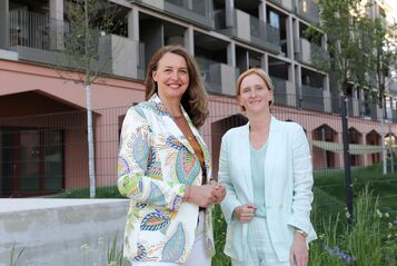 Zwei Frauen stehen vor einem modernen Wohngebäude, eine trägt einen gemusterten Blazer, die andere einen hellen Anzug
