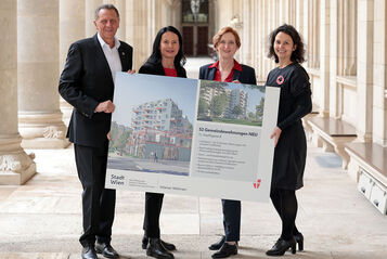 Drei weibliche und eine männliche Person stehen in den Arkaden des Wiener Rathauses und halten gemeinsam ein Schild auf dem Visualisierungen des Gemeindebau NEU zu sehen sind, sowie ein wenig Text