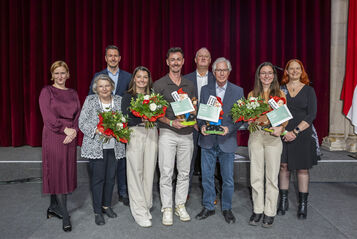 Gruppe von acht Personen vor rotem Vorhang und österreichischer Flagge, drei Personen halten Blumensträuße und Urkunden