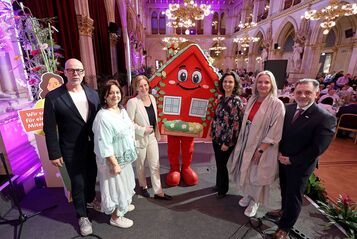 Gruppe von neun Personen steht in festlich beleuchtetem Saal neben wienwohntbesser-Maskottchen, ein rotes Haus mit Gesicht und Blumenschmuck, sie halten ein Schild mit Text 'Wir sorgen für ein gutes Miteinander'