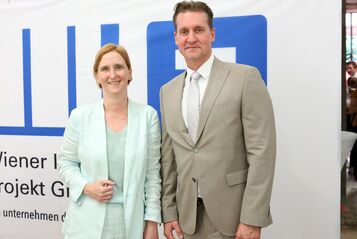 Zwei Personen in Anzügen stehen vor einem weißen Banner mit blauem Logo