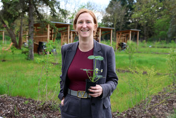 Person mit kurzem Haar und grauem Anzug hält einen kleinen Blumentopf mit einer Pflanze in einem Garten