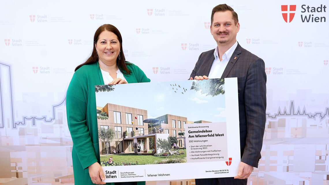 Eine Frau und ein Mann halten ein Schild mit der Visualisierung des neuen Gemeindebaus NEU in der Siedlung Wienerfeld West, erkennbar an der Beschriftung des Schildes. Sie lächeln in die Kamera.