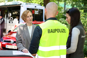 Drei Personen im Gespräch an einem Stehtisch, eine Frau trägt Blazer, ein Mann eine gelbe Weste mit Aufschrift "BAUPOLIZEI", eine weitere Frau trägt schwarze Weste