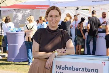 Frau lehnt an einem Schild mit der Aufschrift 'Wohnberatung Wien' auf einer Freiluftveranstaltung mit mehreren Menschen und Informationsständen im Hintergrund
