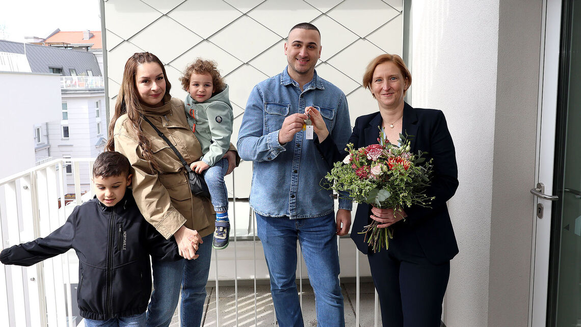 Eine Familie mit 2 kleinen Kindern steht neben einer Person, die einen Blumenstrauß in der Hand hält. Die weibliche Person der Familie hält ein Kind auf dem Arm und das zweite an der Hand, der Vater bekommt den Schlüssel von der Frau mit dem Blumenstrauß gereicht.