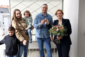Eine Familie mit 2 kleinen Kindern steht neben einer Person, die einen Blumenstrauß in der Hand hält. Die weibliche Person der Familie hält ein Kind auf dem Arm und das zweite an der Hand, der Vater bekommt den Schlüssel von der Frau mit dem Blumenstrauß gereicht.