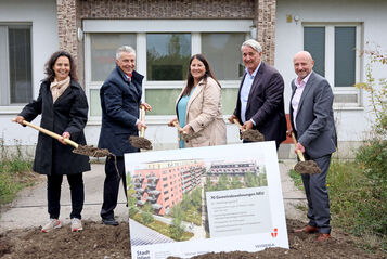 Katharina Klement (Vizedirektorin von Wiener Wohnen), Gemeinderat Josef Taucher, Vizebürgermeisterin und Wohnbaustadträtin Kathrin Gaál, Bezirksvorsteher Ernst Nevrivy und Paul Steurer, Direktor der WIGEBA, stehen jeweils mit einem Spaten voll Erde hinter einer Tafel mit der Visualisierung des Gemeindebau NEU in der Weidingergasse. Sie sind durchwegs herbstlich gekleidet und lächeln in die Kamera. 