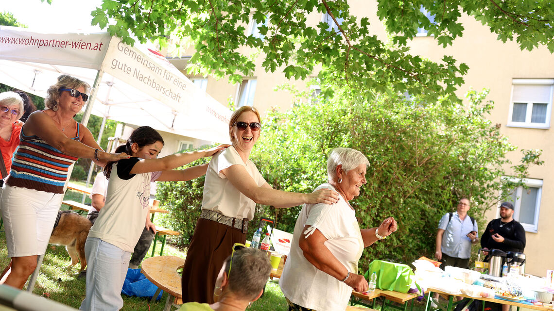 Mehrere Menschen tanzen in einer Reihe im Freien unter Bäumen bei einem Fest mit Tischen und Zelten im Hintergrund