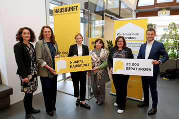 Fünf Personen stehen in einem modernen Innenraum und halten gelbe Schilder mit den Aufschriften '6 JAHRE HAUSKUNFT' und '23.000 BERATUNGEN', im Hintergrund gelbe Banner mit dem Schriftzug 'HAUSKUNFT'