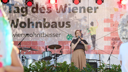 Frau mit Mikrofon auf Bühne vor Banner mit Schriftzug 'Tag des Wiener Wohnbaus' und Schlagzeug im Hintergrund