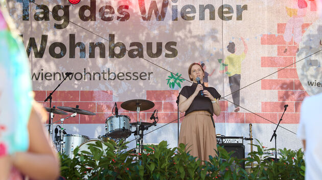 Frau mit Mikrofon auf Bühne vor Banner mit Schriftzug 'Tag des Wiener Wohnbaus' und Schlagzeug im Hintergrund