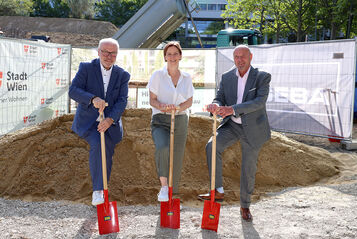 Drei Personen in formeller Kleidung stehen auf einer Baustelle und halten jeweils einen roten Spaten in der Hand, hinter ihnen ein großer Erdhaufen und Bauzäune mit dem Logo der Stadt Wien