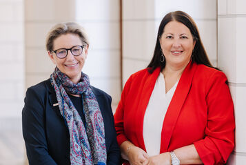 Zwei Frauen, Karin Ramser und Kathrin Gaál, stehen in einem Gang des Rathauses und lächeln in die Kamera. Beide sind businessmäßig mit Blazern gekleidet.