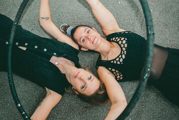 Zwei junge Frauen liegen Kopf neben Kopf in gegengesetzter Richtung auf dem Asphalt und blicken in die Kamera. Sie sind mit schwarzen Shirts und schwarzen Leggings bekleidet und halten einen Hulahoop-Ring in ihren Händen.
